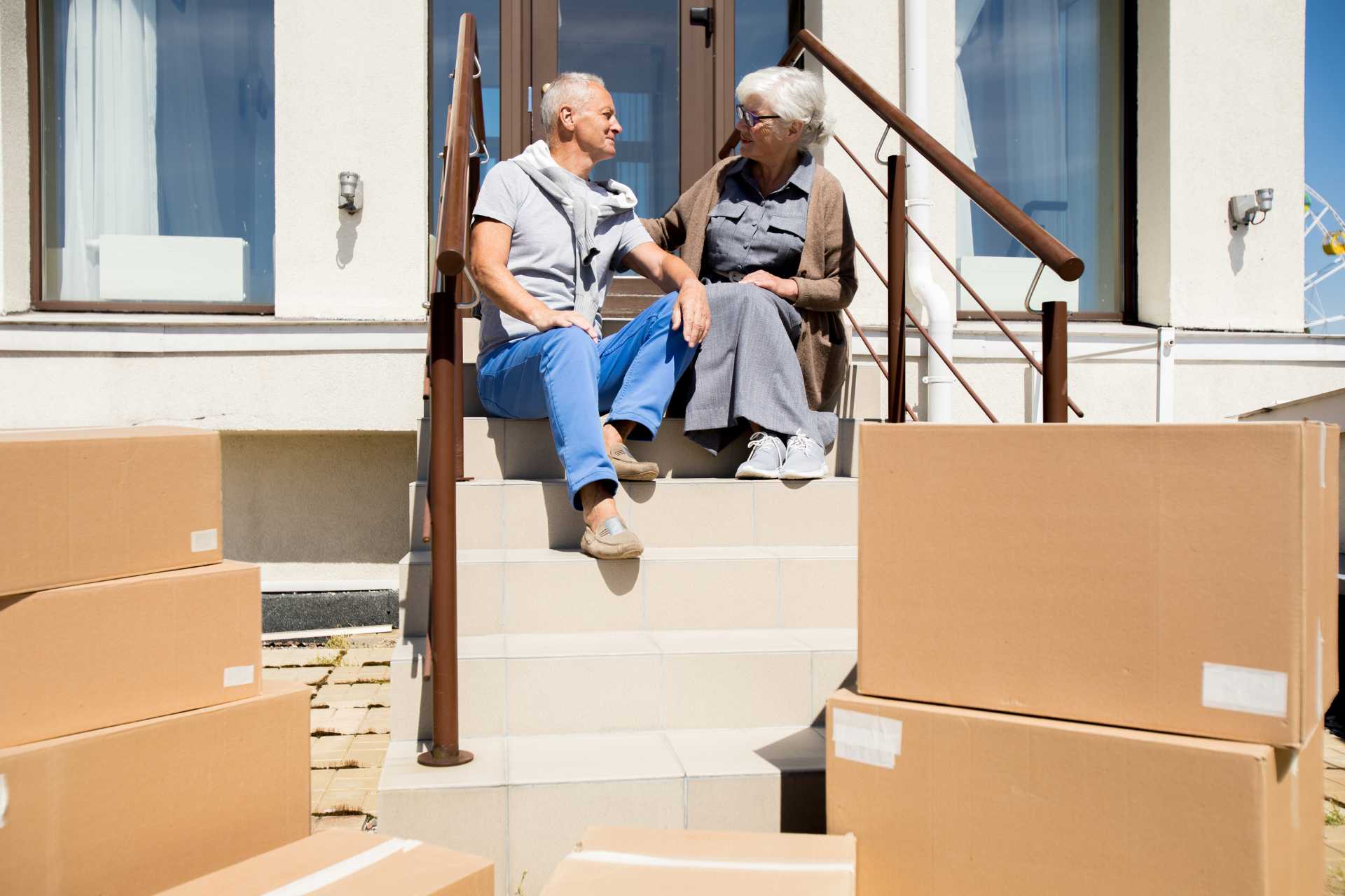 Two smiling seniors sitting on their front porch stoop surrounded by moving boxes.
